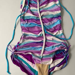May include: A one-piece swimsuit with horizontal stripes in shades of purple, blue, and white. The swimsuit has turquoise drawstrings and a beige lining. The design features a ruched effect and a zipper down the front.