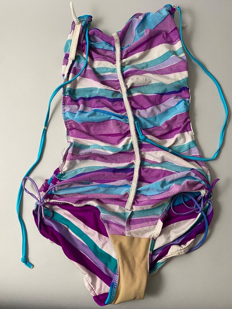 May include: A one-piece swimsuit with horizontal stripes in shades of purple, blue, and white. The swimsuit has turquoise drawstrings and a beige lining. The design features a ruched effect and a zipper down the front.
