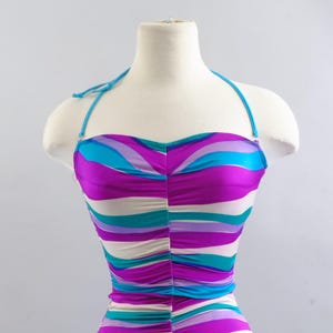 May include: A halter-style swimsuit with a vibrant, striped pattern in shades of purple, turquoise, and white. The swimsuit is displayed on a mannequin, showcasing its design and fit. The top features a ruched detail.