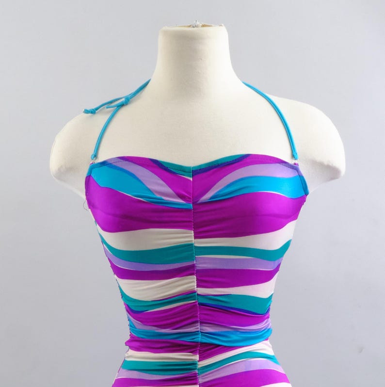May include: A halter-style swimsuit with a vibrant, striped pattern in shades of purple, turquoise, and white. The swimsuit is displayed on a mannequin, showcasing its design and fit. The top features a ruched detail.