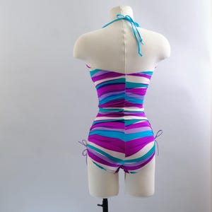 May include: A vintage one-piece swimsuit on a mannequin. The swimsuit features horizontal stripes in shades of purple, turquoise, and white. It has a halter neck and adjustable ties at the sides of the shorts. The fabric is ruched at the waist.