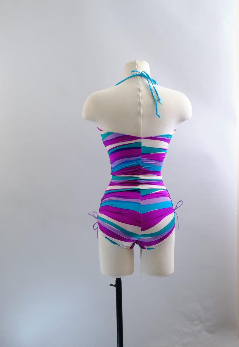 May include: A vintage one-piece swimsuit on a mannequin. The swimsuit features horizontal stripes in shades of purple, turquoise, and white. It has a halter neck and adjustable ties at the sides of the shorts. The fabric is ruched at the waist.