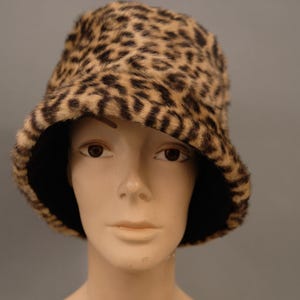 Puede incluir: Un sombrero de cubo con estampado de leopardo y un interior negro. El sombrero presenta un patrón de manchas marrones y negras sobre un fondo beige. El sombrero se muestra en una cabeza de maniquí.