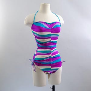 May include: A vintage one-piece swimsuit with a halter neck and a vibrant striped pattern in shades of purple, teal, and cream. The swimsuit is displayed on a mannequin, showcasing its form-fitting design and ruched detailing. The swimsuit has adjustable side ties.
