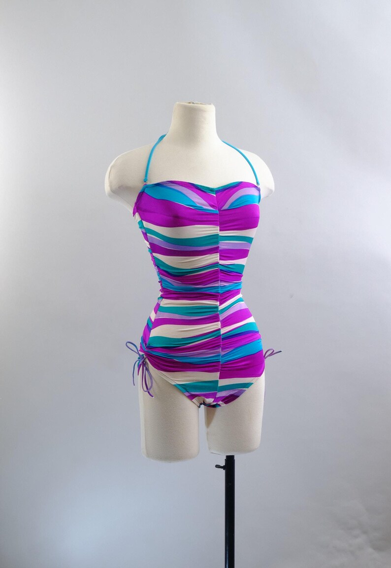 May include: A vintage one-piece swimsuit with a halter neck and a vibrant striped pattern in shades of purple, teal, and cream. The swimsuit is displayed on a mannequin, showcasing its form-fitting design and ruched detailing. The swimsuit has adjustable side ties.