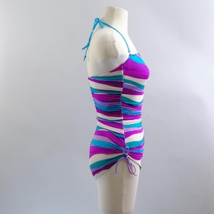 May include: A colorful, striped swimsuit on a mannequin. The one-piece swimsuit features horizontal stripes in shades of purple, teal, and white. It has a halter neck and a ruched side detail. The swimsuit is displayed on a mannequin against a plain background.