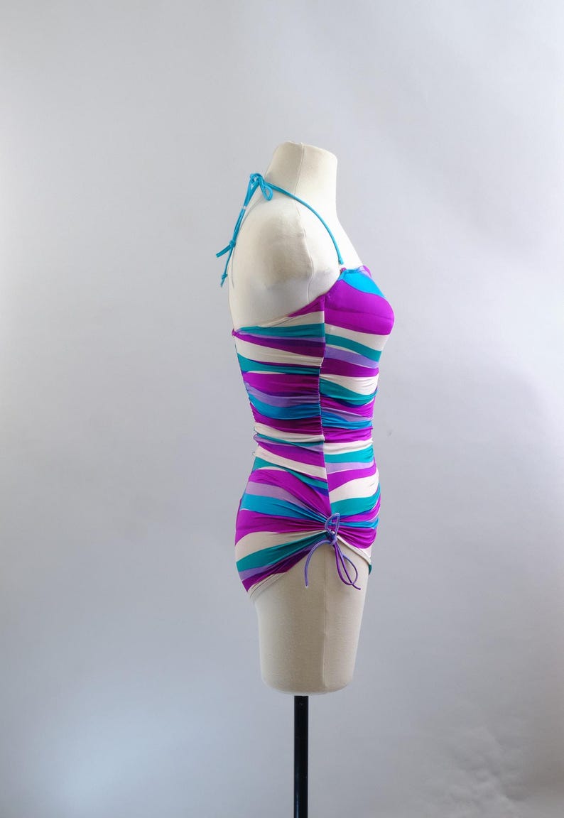 May include: A colorful, striped swimsuit on a mannequin. The one-piece swimsuit features horizontal stripes in shades of purple, teal, and white. It has a halter neck and a ruched side detail. The swimsuit is displayed on a mannequin against a plain background.