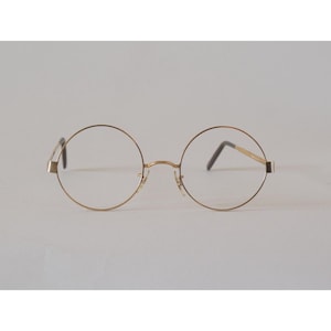 May include: Gold-rimmed round eyeglasses with thin frames. The glasses have a classic, minimalist design, featuring a bridge and temples. The lenses are clear, and the overall aesthetic is vintage-inspired.