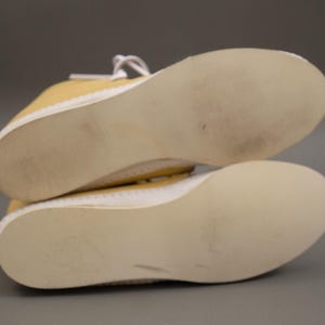 Pu&ograve; includere: Un paio di scarpe da ginnastica gialle in tela con suole e lacci bianchi. Le suole sono beige chiaro e mostrano segni di usura. Le scarpe sono posizionate con le suole rivolte verso l'osservatore.