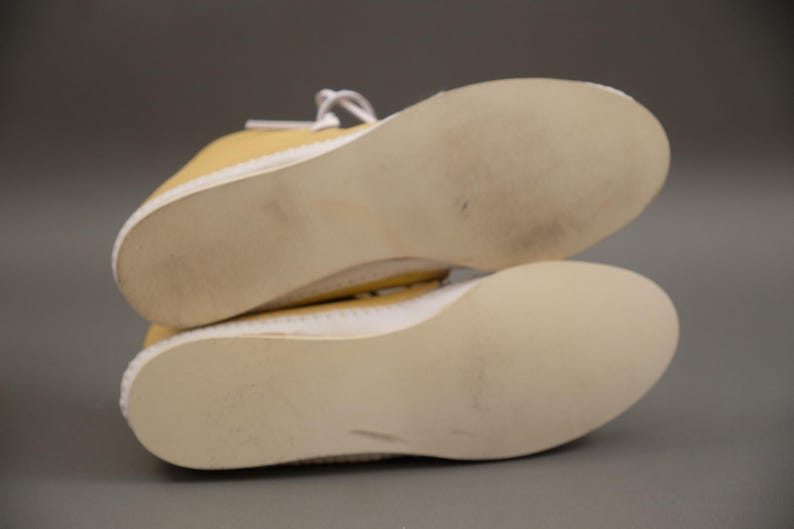 Pu&ograve; includere: Un paio di scarpe da ginnastica gialle in tela con suole e lacci bianchi. Le suole sono beige chiaro e mostrano segni di usura. Le scarpe sono posizionate con le suole rivolte verso l'osservatore.
