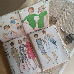 May include: A collection of vintage sewing patterns featuring various dress designs. The patterns include a green dress with a white collar, a red dress with a white collar, a blue dress with a white collar, and a white dress with a blue collar. The patterns are printed on paper and have instructions for sewing the dresses.