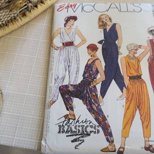 May include: A vintage sewing pattern by McCall's, featuring illustrations of jumpsuits in various colors and styles. The pattern packet includes the text "Fashion Basics" and "Cut-to-Fit". A silver shoe-shaped pin cushion with a red pad and a spool of thread are also visible.