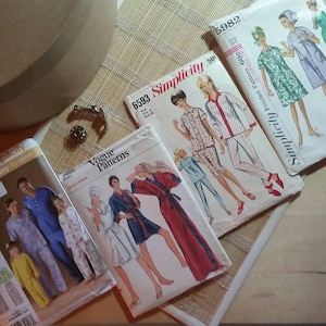 May include: A collection of vintage sewing patterns for clothing, including Simplicity patterns for pajamas and a Vogue pattern for a robe. The patterns feature illustrations of women wearing the garments.