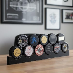 May include: A black wooden display stand holding eleven circular hockey pucks arranged in two rows. The pucks feature various colorful team logos, text including "1st GOAL" and "Bandits," and graphic illustrations of hockey players on their faces.