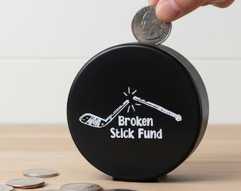 May include: A black circular coin bank shaped like a hockey puck with white text that reads Broken Stick Fund and a graphic of a snapped hockey stick. The bank features a slot at the top for inserting coins, including a silver US quarter coin.
