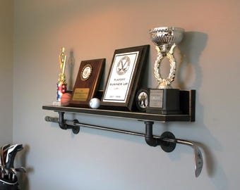 Puede incluir: Un estante de pared negro con una estructura de soporte estilo tubería que integra un palo de golf en el diseño. El estante sostiene varios trofeos deportivos, un balón de baloncesto, una pelota de golf y una placa enmarcada Playoff Runner Up.