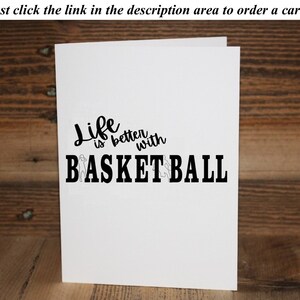 May include: A white card with black text that reads "Life is better with BASKETBALL". The word "Life" is written in a cursive font. The word "BASKETBALL" is in a bold, block font. There are two small silhouettes of basketball players on either side of the word "BASKETBALL".