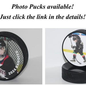 May include: Two hockey pucks with personalized photos and the text "BANDITS 2024" printed on them. The pucks are on a black base.