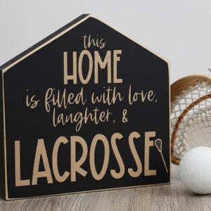 May include: A house-shaped black wooden sign with tan lettering that reads: this HOME is filled with love, laughter, & LACROSSE, featuring a small lacrosse stick icon. Next to the sign is a white lacrosse ball and a wooden lacrosse stick head.
