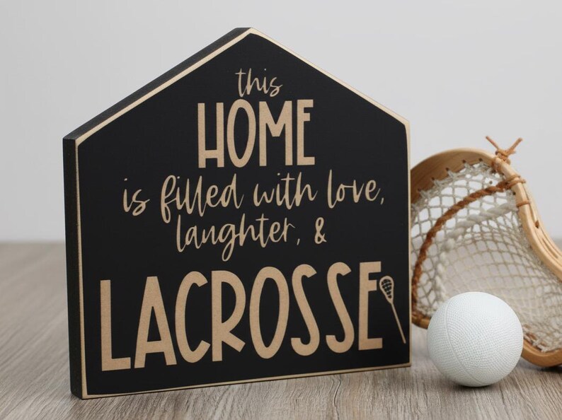 May include: A house-shaped black wooden sign with tan lettering that reads: this HOME is filled with love, laughter, & LACROSSE, featuring a small lacrosse stick icon. Next to the sign is a white lacrosse ball and a wooden lacrosse stick head.