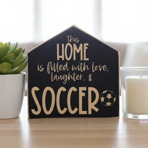 May include: A house-shaped black wooden sign with light-colored text that reads: this HOME is filled with love, laughter, & SOCCER, featuring a small graphic of a soccer ball next to the word SOCCER, with a clean, flat, and matte finish.