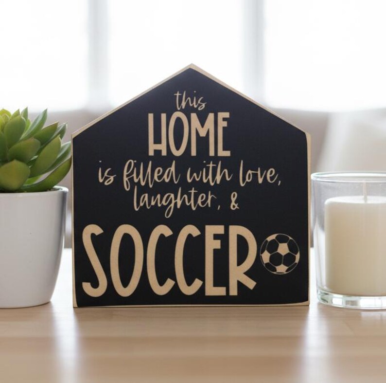 May include: A house-shaped black wooden sign with light-colored text that reads: this HOME is filled with love, laughter, & SOCCER, featuring a small graphic of a soccer ball next to the word SOCCER, with a clean, flat, and matte finish.