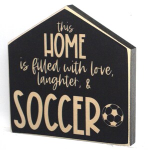 Op de afbeelding: Zwart houten bord in de vorm van een huis. Het bord heeft de tekst "This home is filled with love, laughter, & soccer" met een voetbal afbeelding.