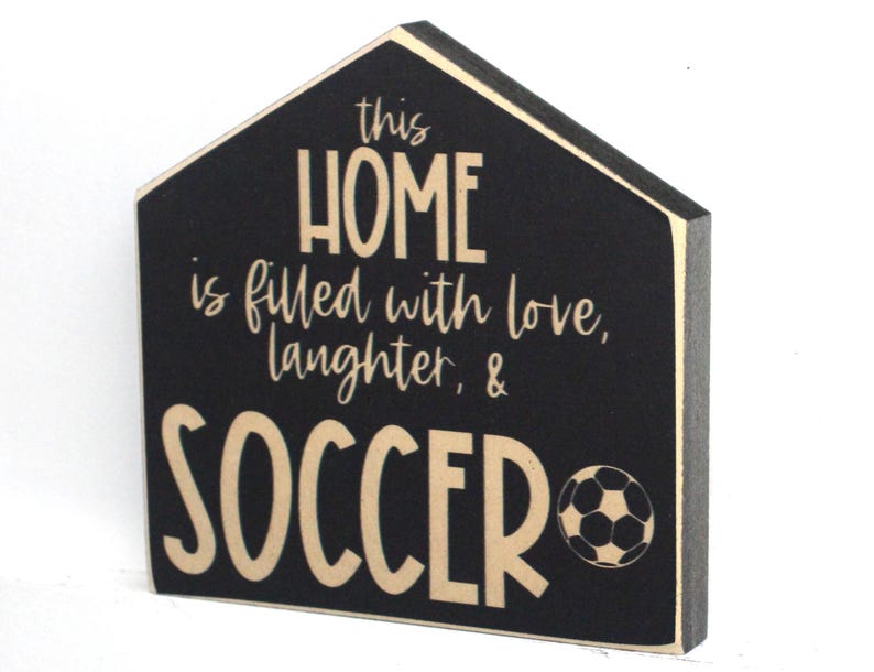 Op de afbeelding: Zwart houten bord in de vorm van een huis. Het bord heeft de tekst "This home is filled with love, laughter, & soccer" met een voetbal afbeelding.