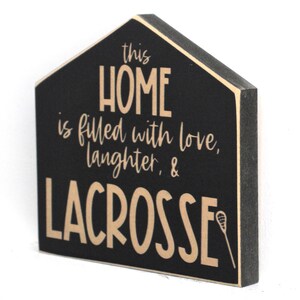 Op de afbeelding: Een zwart houten bord in de vorm van een huis. Het bord heeft de tekst "This home is filled with love, laughter, & lacrosse".