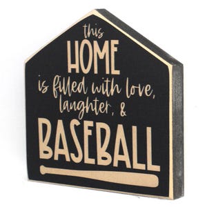 Op de afbeelding: Een zwart houten bord in de vorm van een huis met de tekst "This home is filled with love, laughter, & baseball" in beige letters. Een beige honkbalknuppel is afgebeeld onder de tekst.