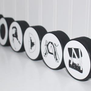 May include: A set of seven black hockey pucks with white designs. Each puck features a different hockey-related image, including a hockey stick, a hockey player, and a hockey puck.