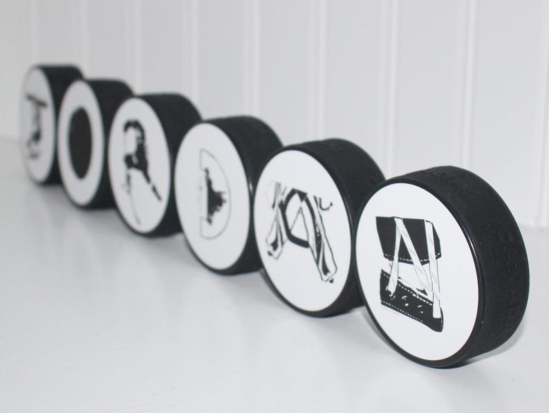 May include: A set of seven black hockey pucks with white designs. Each puck features a different hockey-related image, including a hockey stick, a hockey player, and a hockey puck.