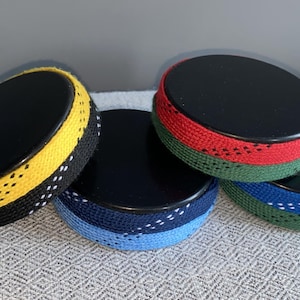 May include: Four black, round objects with colorful trim. One has yellow trim, another has red and green trim, and a third has blue trim. The trim appears to be made of a woven material. The objects are arranged on a textured, light gray surface.