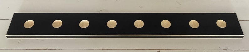 May include: A rectangular black wooden coat rack with seven evenly spaced, gold-colored knobs. The rack has a thin gold trim along the bottom edge and is mounted on a white wooden surface.