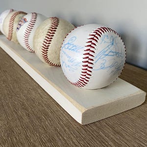 May include: A display of four baseballs with red stitching and white surfaces, resting on a light-colored wooden shelf. The baseballs have blue signatures. The shelf is rectangular with a white painted edge.