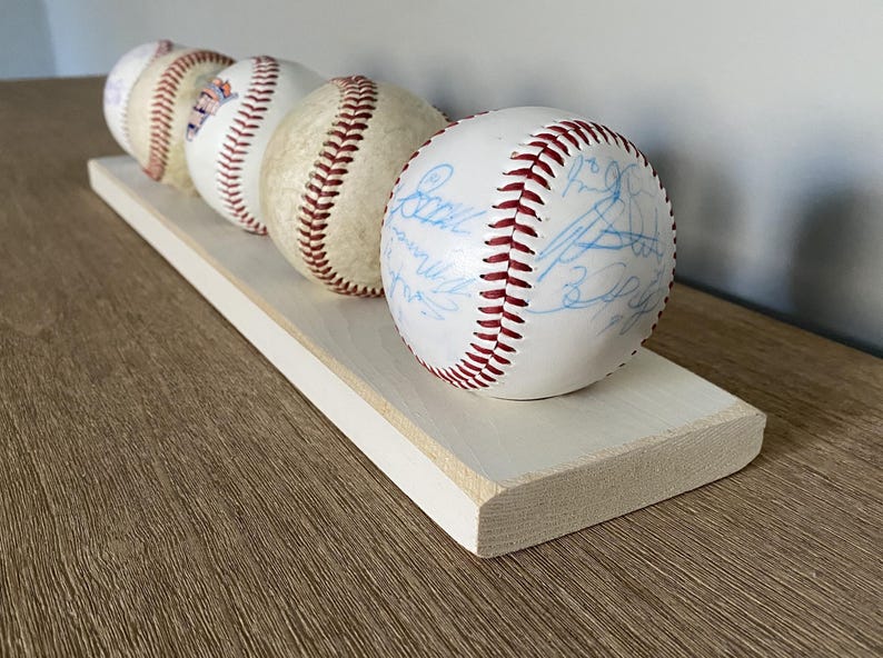 May include: A display of four baseballs with red stitching and white surfaces, resting on a light-colored wooden shelf. The baseballs have blue signatures. The shelf is rectangular with a white painted edge.