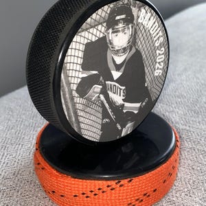 May include: A black hockey puck with a black and white image of a hockey player in a net. The puck is mounted on a second puck with orange tape around the edge. The text "Bandits 2026" is visible on the puck.