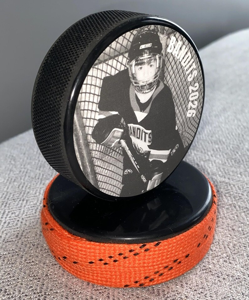 May include: A black hockey puck with a black and white image of a hockey player in a net. The puck is mounted on a second puck with orange tape around the edge. The text "Bandits 2026" is visible on the puck.