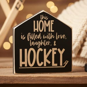 A black wooden sign in the shape of a house with the text "This Home is filled with love, laughter & HOCKEY" in engraved lettering. The sign is sitting on a table with a puck and twinkling lights in the background.