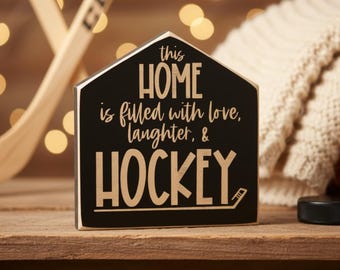 May include: A house-shaped wooden sign with a black finish and light-colored edges. The text reads: this HOME is filled with love, laughter, & HOCKEY. The word HOCKEY is underlined with a graphic of a hockey stick blade at the end of the line.