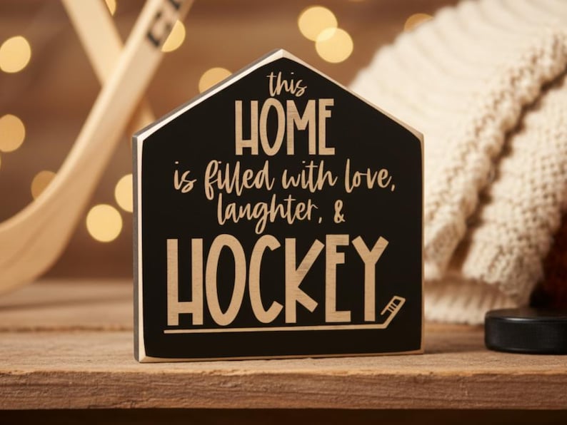 A black wooden sign in the shape of a house with the text "This Home is filled with love, laughter & HOCKEY" in engraved lettering. The sign is sitting on a table with a puck and twinkling lights in the background.