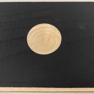 May include: A round, light-colored wooden disc is centered on a black surface. The disc has a visible wood grain pattern. The background is a solid black color, with a lighter border at the top and bottom.