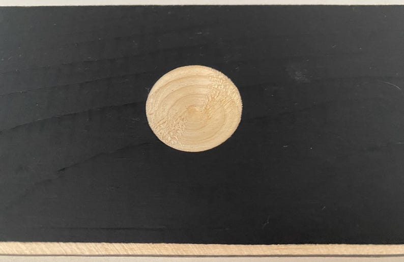 May include: A round, light-colored wooden disc is centered on a black surface. The disc has a visible wood grain pattern. The background is a solid black color, with a lighter border at the top and bottom.