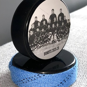 May include: A black hockey puck with a team photo and the text "Bandits 2025-26" on top, resting on a second puck wrapped with blue hockey tape. The pucks are on a light gray surface.