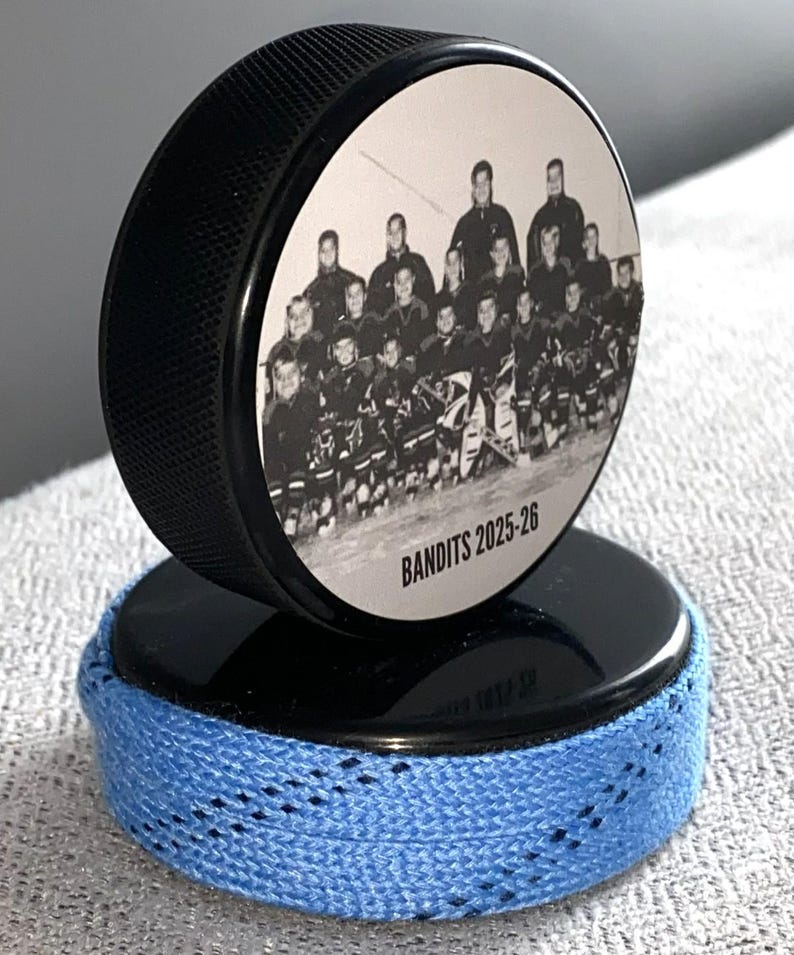 May include: A black hockey puck with a team photo and the text "Bandits 2025-26" on top, resting on a second puck wrapped with blue hockey tape. The pucks are on a light gray surface.