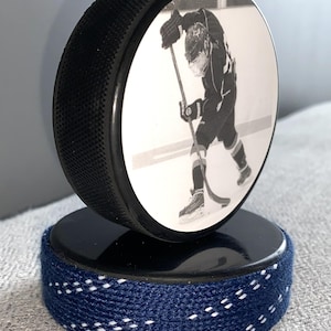 May include: A black hockey puck with a black and white image of a hockey player on the front. The puck sits atop a second puck wrapped in navy blue fabric with white stitching.