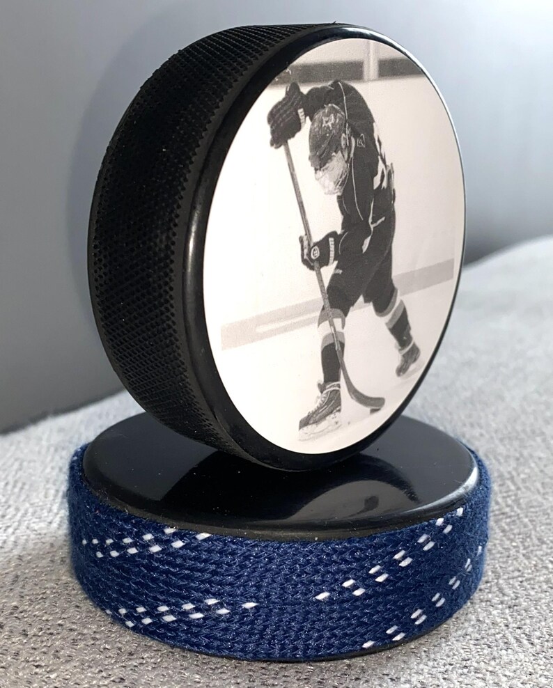 May include: A black hockey puck with a black and white image of a hockey player on the front. The puck sits atop a second puck wrapped in navy blue fabric with white stitching.