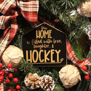 Op de afbeelding: Een zwart en gouden bord met de tekst "This home is filled with love, laughter, & hockey" hangt aan een kerstkrans met rode bessen, geruit lint en dennentakken.