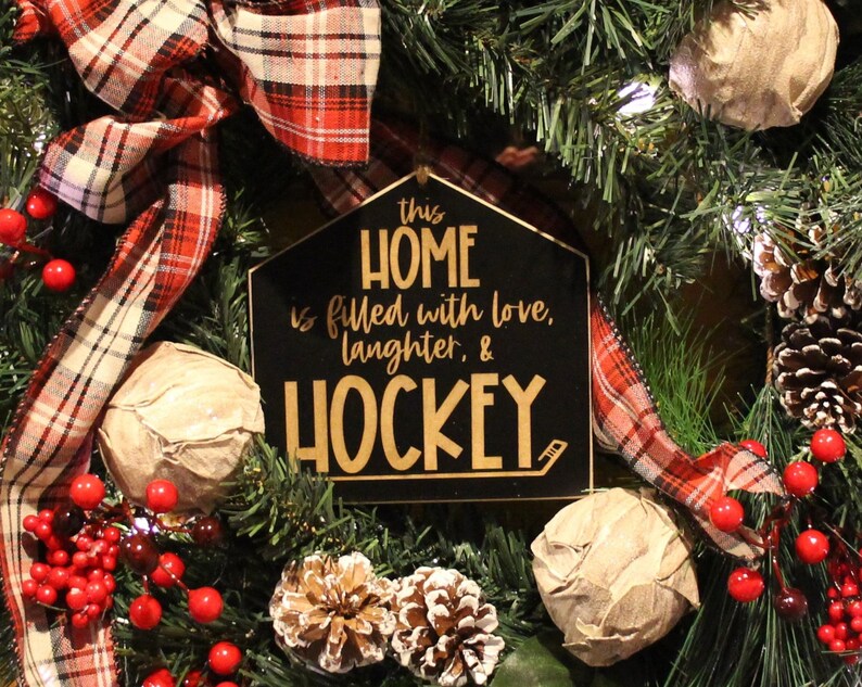 Op de afbeelding: Een zwart en gouden bord met de tekst "This home is filled with love, laughter, & hockey" hangt aan een kerstkrans met rode bessen, geruit lint en dennentakken.