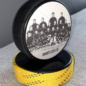 May include: A black hockey puck with a team photo and the text "BANDITS 2025-26" on a black base with yellow and black trim. The puck is standing upright, showcasing the team photo.